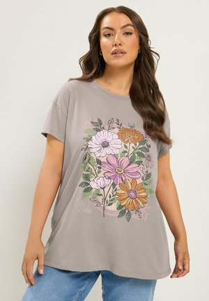 Kobieta ubrana w luźny szary t-shirt z dużym kwiatowym nadrukiem w różowym, pomarańczowym i zielonym kolorze, w połączeniu z niebieskimi dżinsami, stojąca na jednolitym tle.