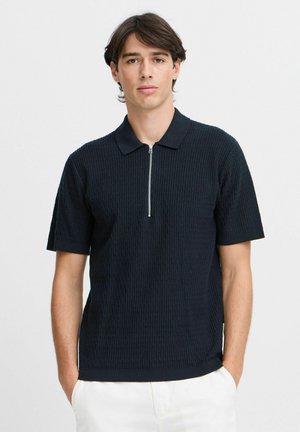Polo à manches courtes bleu marine avec un design en maille texturée, présentant un col zippé et des poignets côtelés. Tissu lisse, coupe ajustée.