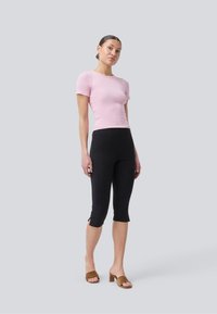 Helles rosa, geripptes T-Shirt mit kurzen Ärmeln, kombiniert mit schwarzen, knöchellangen Leggings mit seitlichen Schlitzen. Das Modell trägt braune Sandalen mit Blockabsätzen.