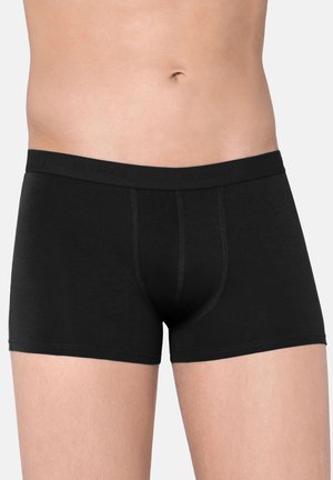 Mies vartalo mustat tiukat bokserit jalassa seisoo yksiväristä taustaa vasten.