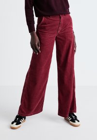 Pantaloni din catifea cu picioare largi în nuanța burgundă, având nervuri verticale texturate, buzunare frontale și talie înaltă. Asortați cu teniși negri și crem.