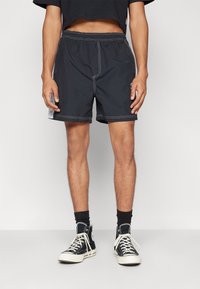 Černé plavecké šortky s elastickým pasem, bílým prošíváním a bočními kapsami, spojené s černými vysokými teniskami a ponožkami.