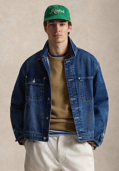 Άνδρας με πράσινο καπέλο που γράφει "Ralph," σκούρο μπλε τζιν μπουφάν, μπεζ φούτερ, ριγέ πουκάμισο από κάτω και ανοιχτόχρωμο παντελόνι, με τα χέρια στις τσέπες.