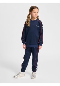 Marinblå träningströja med röda detaljer och logotyp, ihopparad med matchande joggingsbyxor med liknande detaljer. Silverfärgade skor fulländar looken.