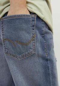 Denim jeans v bledi modri barvi, s hrbtnim žepom z rumenim šivom in značilnim ukrivljenim dizajnom.
