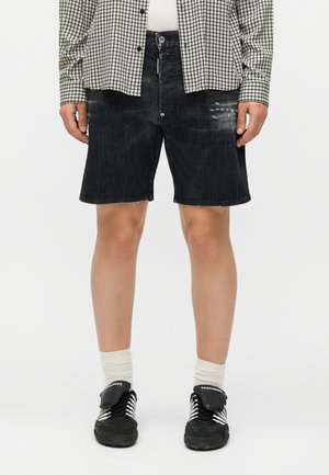 Άτομο που φοράει μαύρα τζιν σορτς με ελαφρές φθορές, καρό πουκάμισο με κουμπιά, άσπρες κάλτσες και μαύρα αθλητικά παπούτσια με άσπρα κορδόνια.