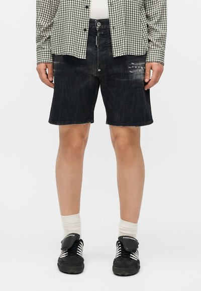 Personne portant un short en jean noir légèrement usé, une chemise à carreaux boutonnée, des chaussettes blanches et des baskets noires avec des lacets blancs.