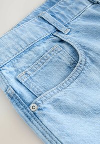 Tejido de denim azul claro con una textura sutil, con un bolsillo frontal y costuras visibles. Incluye un detalle de botón de metal.