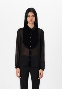 Blusa preta transparente com colarinho de veludo, apresentando fechamento frontal com botões e mangas longas com punhos botão. Cor preta sólida em toda a peça.