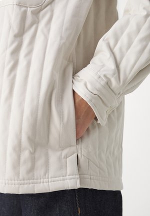 Zbliżenie dłoni osoby w bocznej kieszeni pikowanej, jasnobeżowej kurtki z pionowym przeszyciem, noszonej na ciemnych dżinsach.