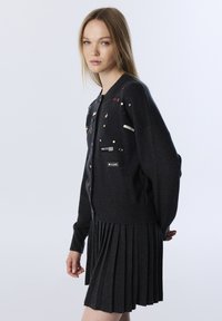 Jonge vrouw in een donkergrijze cardigan met pins en patches, en een bijpassende geplooide rok, staande tegen een effen lichte achtergrond.