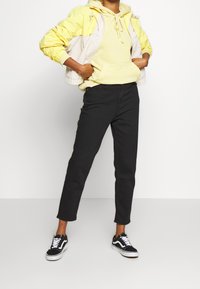 Bunda do deště v žluté a béžové barvě, s kapucí a šňůrkami; spárovaná se světle žlutou mikinou a černými zúženými kalhotami; černé tenisky.