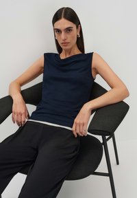 Top senza maniche blu navy con scollo drappeggiato, realizzato in tessuto morbido, abbinato a pantaloni neri larghi, seduto su una moderna sedia nera.