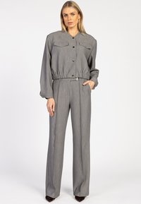 Vrouw die staat in een grijze knopenjasje tot de taille met klepzakken en bijpassende hoogmatige wijde broek, met de handen deels in de zakken.