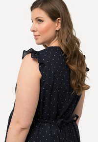 Abito blu con pois bianchi, caratterizzato da maniche a volants e una vita legata. Tessuto leggero con una texture liscia.