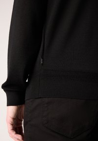 Pull en maille noir avec des poignets et un ourlet côtelés ; petite étiquette sur le côté. Porté avec un pantalon noir, mettant en valeur la texture lisse du tissu.