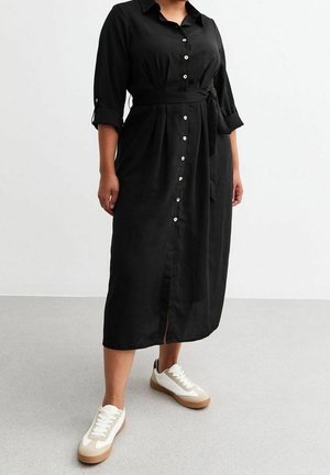 Vrouw die een zwart midi-jurk met knopen draagt, met opgerolde mouwen en een taillekoord, gecombineerd met witte en beige sneakers, staand op een grijze vloer.