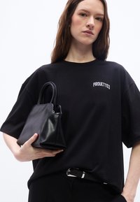 Νεαρή γυναίκα με μακριά καστανά μαλλιά που φορά μαύρο μπλουζάκι «Pirouettes», κρατώντας μια μικρή μαύρη δερμάτινη τσάντα σε λευκό φόντο.