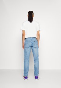 Person med långt hår uppsatt i en hästsvans, klädd i en vit t-shirt, ljusblå jeans och lila samt gula sneakers, stående mot en slät vägg.