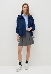 Chaqueta de lluvia impermeable en color azul marino con capucha, falda plisada gris, camisa azul, zapatillas deportivas blancas y calcetines negros, con un fondo neutro.