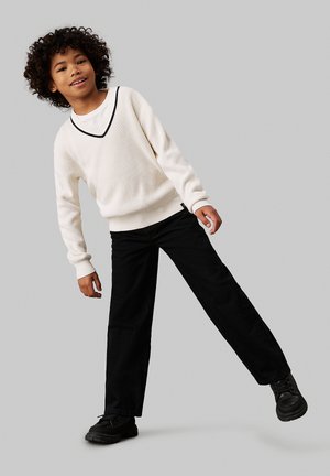 Suéter blanco de canalé con un ribete en pico negro, combinado con pantalones negros de pierna ancha y zapatos negros. Cabello rizado, fondo neutro.