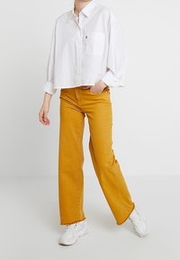 Camisa blanca corta con botones y bolsillo en el pecho, combinada con jeans acampanados color mostaza. Calzado: zapatillas blancas con suelas texturizadas.