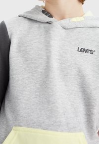 Bambino che indossa un felpa grigia con cappuccio di Levi's, con maniche nere e una tasca anteriore di un lieve giallo pallido, tagliata a livello del collo e del torso.