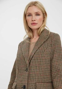 Hnědý kostkovaný blazer s vykrojeným límcem, dvěma knoflíky a kapsovými detaily. Má strukturovanou tkaninu s kontrastními vzory.