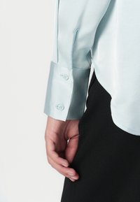 Lys blå satin skjorte med en glatt tekstur, med spiss krage og knapp på mansjetten. Svart stoff er synlig ved kanten.