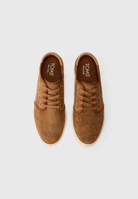 Zapatillas de gamuza marrón con punta redonda, cierre de cordones de seis ojetes y suelas de goma color crema. Presenta un logotipo en la plantilla y perforaciones sutiles.