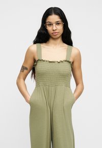 Γυναίκα με μακριά μαύρα μαλλιά και γυαλιά, φοράει αμάνικη ολόσωμη φόρμα σε ελιά πράσινο με βολάν στην κορυφή και τα χέρια στις τσέπες, στέκεται μπροστά σε ένα απλό λευκό φόντο.