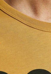 Koszulka bawełniana w musztardowym kolorze z okrągłym dekoltem. Gładka tekstura z ciemnym okrągłym wzorem z przodu. Prosty, casualowy design.