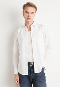 Camisa de algodón blanca de botones con cuello extendido, mangas largas y textura lisa, llevada sobre una camiseta blanca y combinada con jeans oscuros.