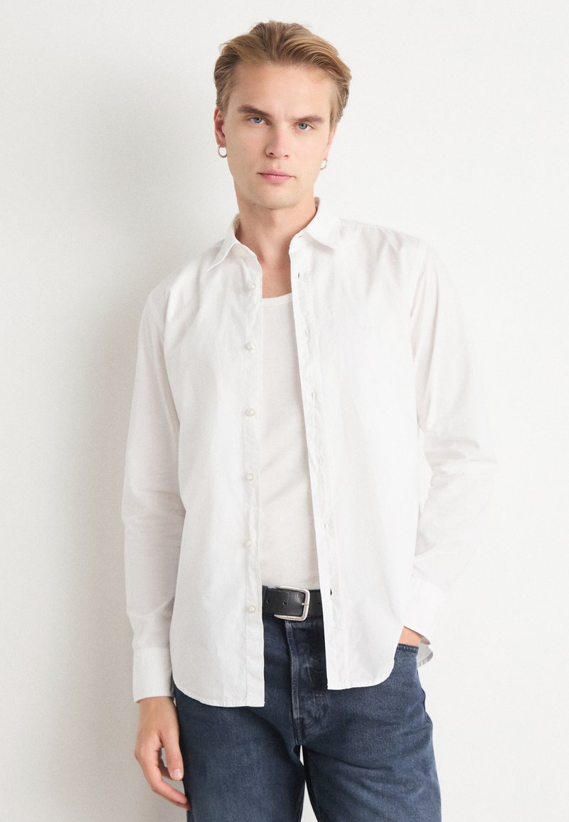 Camisa de algodón blanca de botones con cuello extendido, mangas largas y textura lisa, llevada sobre una camiseta blanca y combinada con jeans oscuros.