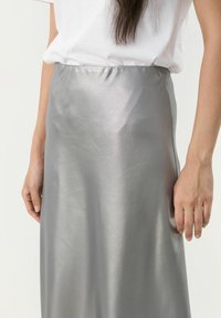 Gonna maxi in satin argento con una superficie liscia e lucida e cintura elastica, abbinata a una t-shirt bianca aderente. Mano sulla cintura.