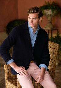 Marinblå stickad cardigan över ljusblå skjorta, matchat med ljusrosa byxor. Sitter på en vävd stol, naturlig miljö med växter i bakgrunden.