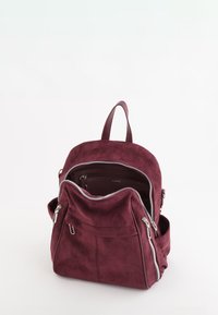Bordeaux ruskindsrygsæk med sølv lynlåse, dobbelte sidelommer og justerbare stropper. Øverste håndtag til bæring. Glat tekstur, afslappet design.