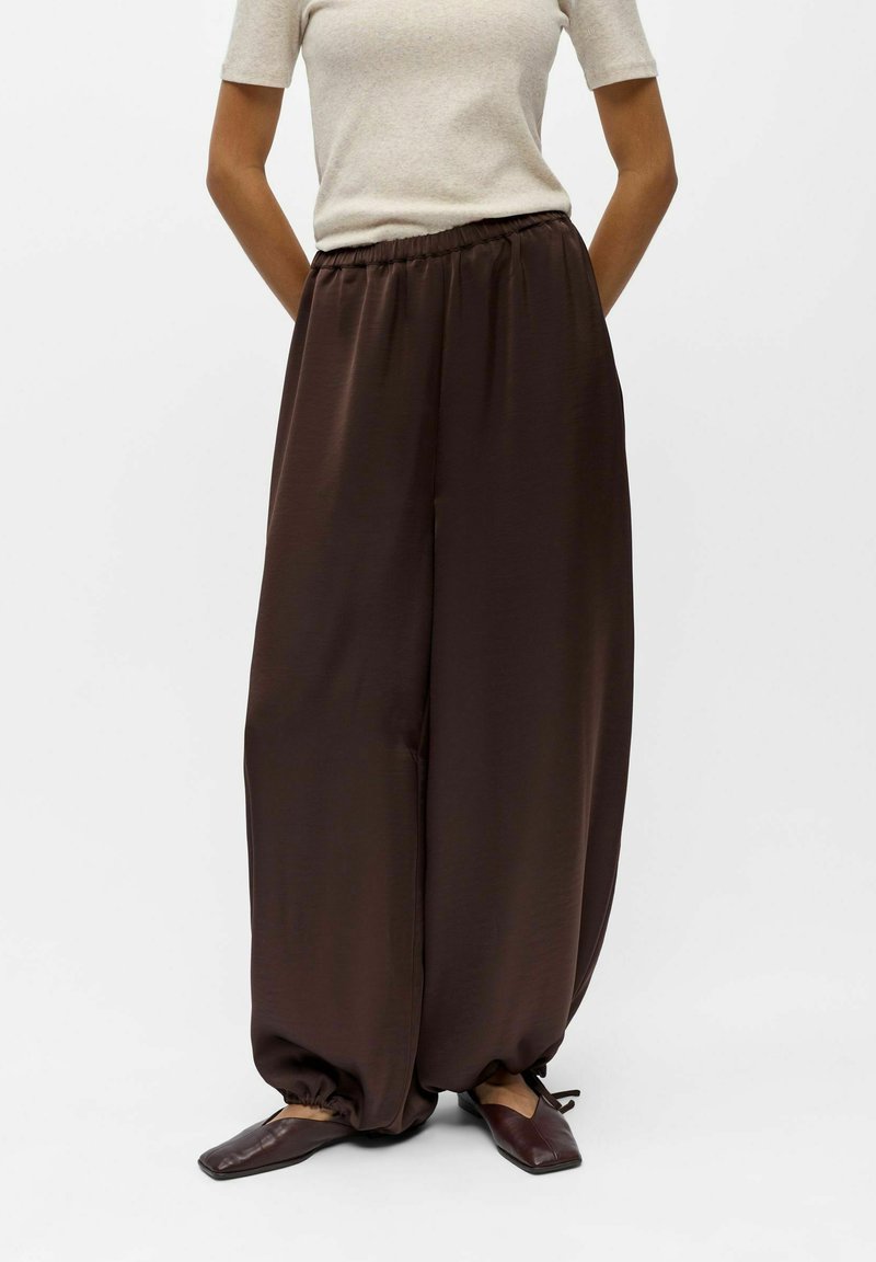Personne portant un pantalon ample marron foncé avec un ourlet froncé, associé à des chaussures marron foncé à bout carré et un haut beige à manches courtes.