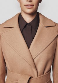 Płaszcz z wełny wielbłądziej z szerokimi klapami i paskiem w talii, noszony na ciemnobrązowej koszuli z guzikami, o gładkiej fakturze i prostych liniach.