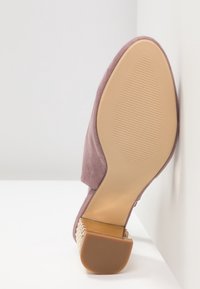Sandale en daim mauve à bout ouvert avec semelle beige texturée et talon bloc bas à motif, positionnée droite contre un mur blanc d’angle.