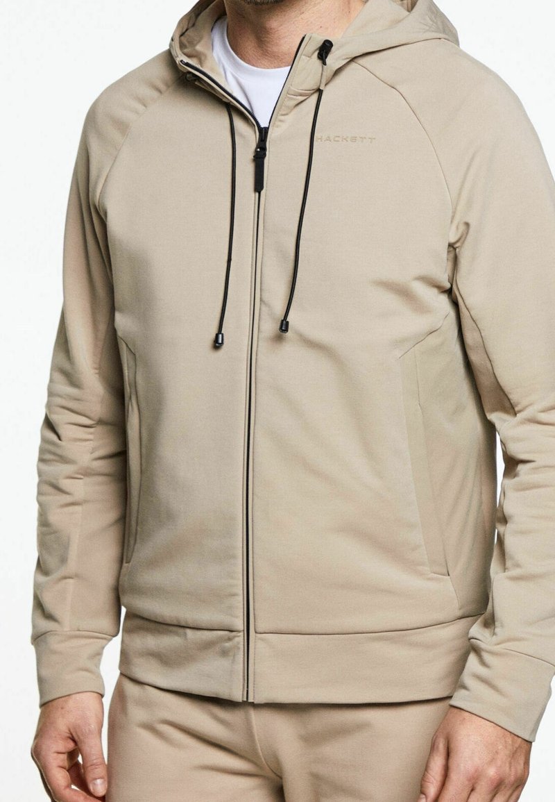 Sudadera beige con cremallera y capucha ajustable, hecha de un tejido suave; cuenta con bolsillos laterales y una textura suave con un diseño ligero.