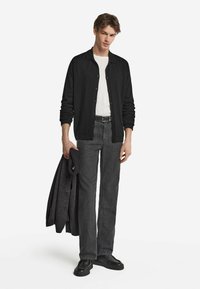 Svart cardigan, vit t-shirt, mörkgrå jeans och svarta skor. Håller en mörkgrå jacka. Avslappnad passform, texturerad tyg och knappr detaljer.