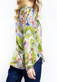 Kvinde iført en langærmet skjorte med et farverigt abstrakt mønster i grøn, lilla, orange og blå, kombineret med mørkeblå jeans.