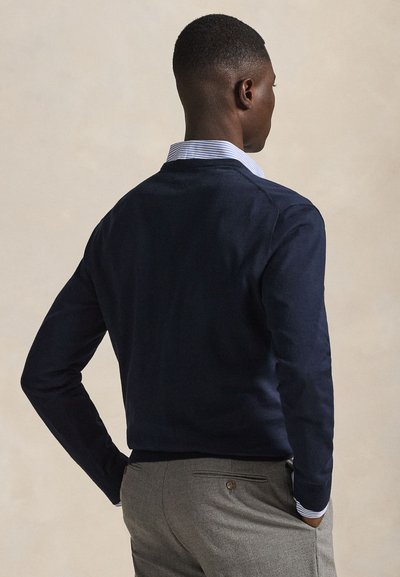 Uomo con capelli corti che indossa un maglione blu navy, una camicia a righe e pantaloni grigi, in piedi con una mano in tasca, rivolto di spalle.