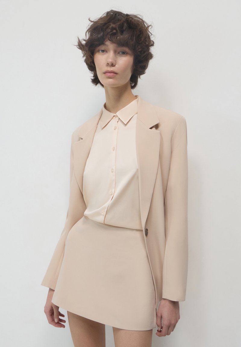 Giovane con i capelli corti e ricci indossa un blazer beige, una camicetta cream con bottoni e una mini gonna asimmetrica coordinata su uno sfondo bianco.