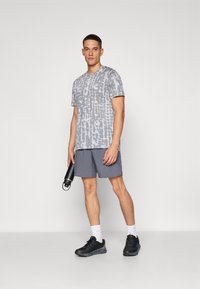 Grå tie-dye kortermet t-skjorte, grå shorts, svarte treningssko, holder en svart vannflaske, mot en enkel bakgrunn.