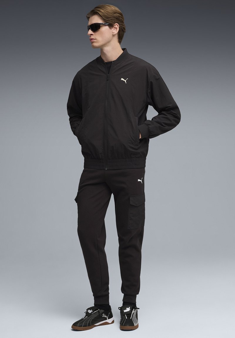 Jeune homme portant un survêtement Puma noir, les mains dans les poches, lunettes de soleil noires, debout devant un fond gris.