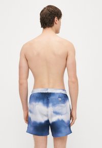 Mėlyni plaukiojimui skirti šortai su dažymo efektu, elastinga liemene, galine kišene ir lengva medžiaga, turintys gradientą nuo tamsiai mėlynos iki šviesiai mėlynos spalvos.