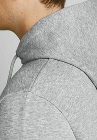 Felpa grigia chiara con cappuccio, caratterizzata da una texture morbida, cuciture delle spalle arrotondate e dettaglio con coulisse. Design semplice senza motivi.