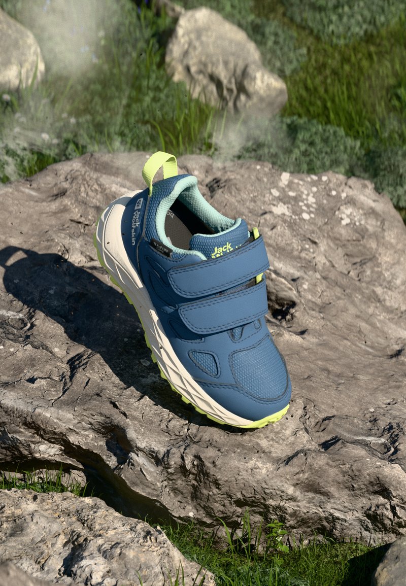 Kék sportcipő tépőzáras pántokkal, textúrált felsővel és világos zöld talppal, egy köves felületen fű között elhelyezve.